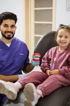 A friendly male dentist in scrubs shows a young girl a dental model and toothbrush in a clinic setting