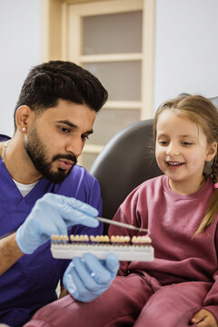 A male dentist in blue scrubs and gloves shows a young girl a shade guide for teeth in a dental clinic setting
