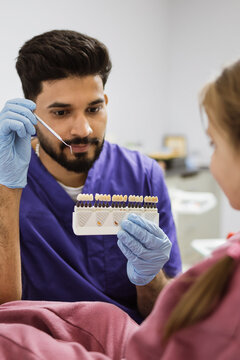 A male dentist in blue gloves holds a dental shade guide while consulting with a patient about tooth color options