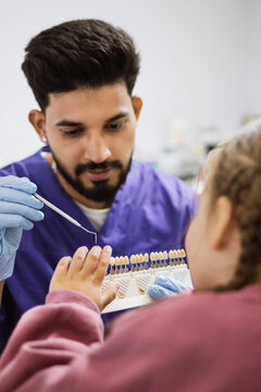 A dentist in a purple uniform and blue gloves holds a dental shade guide and a tool, comparing it to a child's teeth
