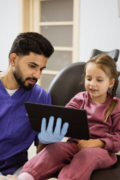 A male dentist in blue scrubs and gloves shows a tablet to a young girl sitting in a dental chair, engaging her with technology during her appointment