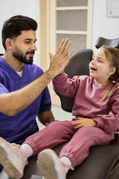 A male dentist in blue scrubs gives a young girl in a pink outfit a high-five while she sits in a dental chair, fostering a positive patient experience