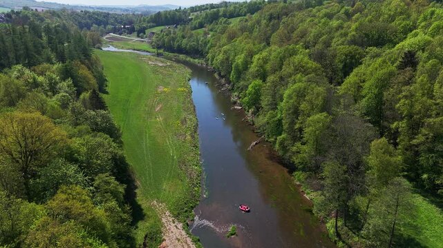 Aerial view of a winding river flowing through a green valley surrounded by dense deciduous forest, with small boats and a raft floating downstream on a sunny day.