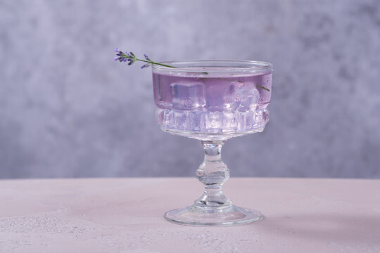 A glass filled with a purple drink sits on a light-colored table. Ice cubes fill the glass, and a sprig of lavender rests on the rim. The background is simple and plain.