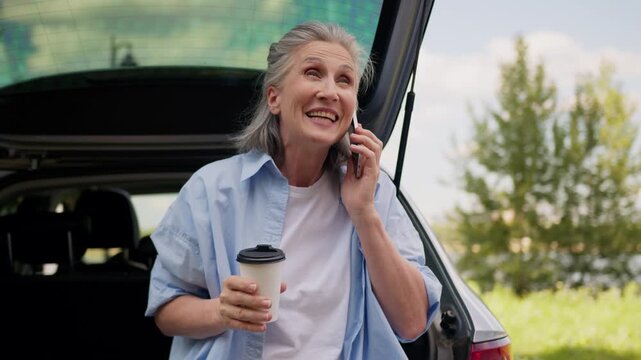 Cheerful Caucasian retired old senior woman calling talking mobile phone with family drinking coffee sitting in car auto trunk at nature travel aged mature senior female talk laugh near automobile