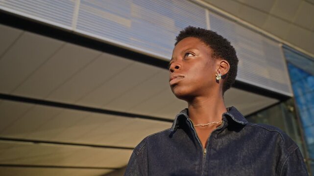 African American young adult woman standing near modern office building, looking up thoughtfully as camera rotates. Footage perfect for corporate diversity and recruitment campaigns