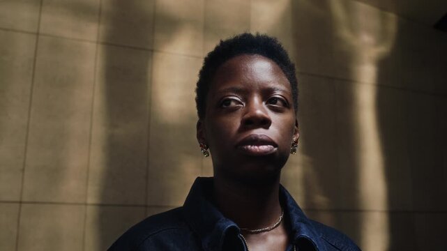 Dolly in close up of Black young adult woman standing in shadowed architectural space, looking around navigating city. Video suitable for social diversity, mental health, or lifestyle campaigns