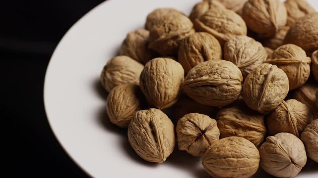 Pile of whole raw walnuts resting on a white plate rotating slowly in a dark moody studio setting
