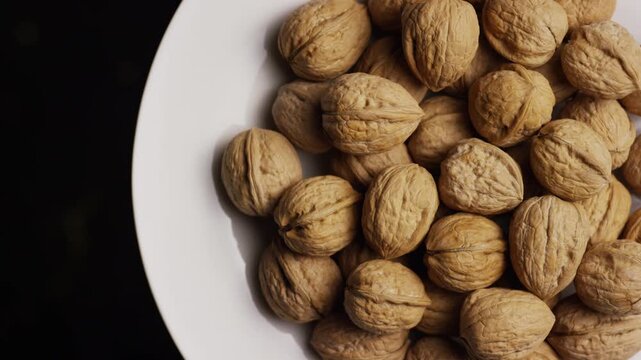 Close up overhead view of a white ceramic bowl filled with organic walnuts rotating on dark background
