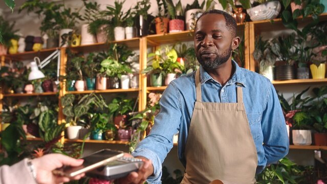 Cheerful positive attractive African American male florist selling bouquets of flowers holding payment terminal at beautiful cozy flower shop. Happy African man smiling enjoying working.