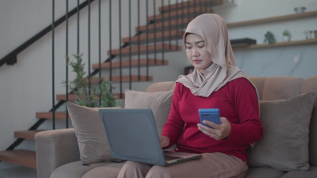 Muslim woman wearing a hijab sits on a living room sofa while using a laptop and smartphone for two-factor authentication, illustrating online security, secure login, and digital banking.