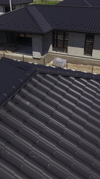 Vertical video. Aerial view moving along the black metal tile roof of a modern house under construction, detailing the material, gutter system, and snow guards on a sunny day with distinct shadows