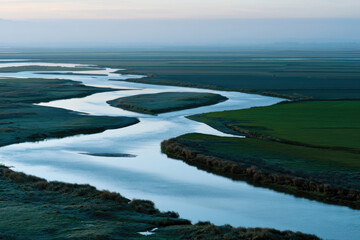 Fototapeta premium A gentle river weaving through lush green fields under a serene sky, providing a picturesque view of nature's beauty and the tranquility of the rural landscape.