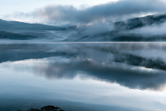 A serene lake enveloped in soft fog casts an enchanting atmosphere over distant hills, creating a mysterious and tranquil landscape at dawn.