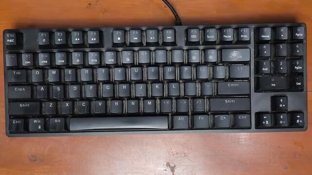 Black keyboard sits on wooden surface. Hand rests near keyboard's right side. Keys are uniform and unlit. Cable trails from back of device. Simple, functional design for typing. Ideal for tech, work