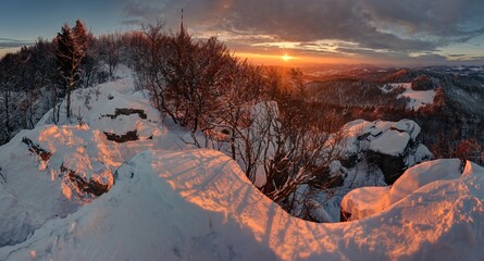 Stunning winter landscape at sunset with snow-covered rocks and trees illuminated by warm golden sunlight. Breathtaking panoramic view of mountains in winter season.