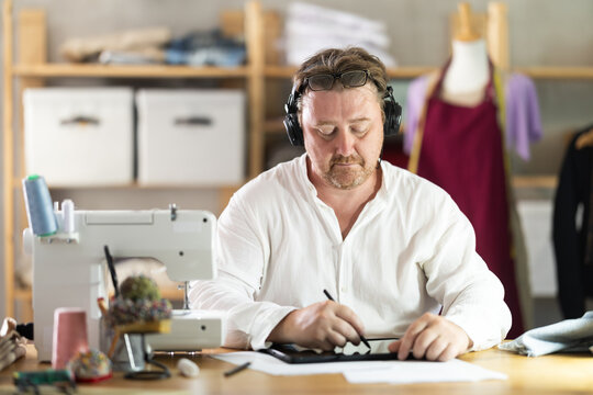 Middle-aged male dressmaker wearing earphones drawing on tablet in sewing workshop