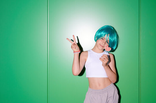 Young girl with vibrant blue wig and lollipop making peace sign against a bright green wall, full of