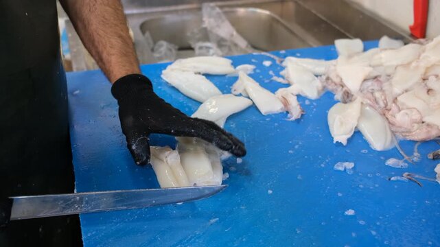 Male hands with black gloves chopping fresh raw squid on a blue cutting board at a fish market, fa01