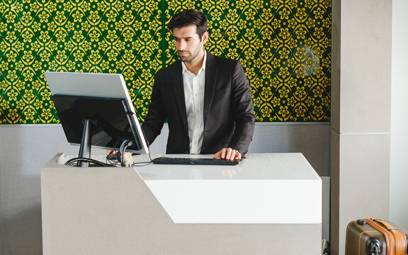 A smartly dressed receptionist works at a modern hotel front desk, using a computer to assist guests; a suitcase rests nearby against a patterned wall.