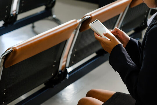 Person seated in a waiting area checks a smartphone beside empty brown metal benches, suggesting an airport or station.