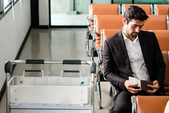 Business traveler in a suit sits in an empty airport lounge, reviewing a passport and boarding pass beside a luggage cart while waiting to board.