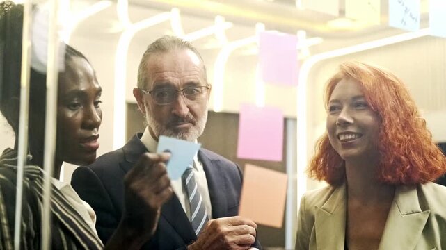Diverse coworkers brainstorm with sticky notes on a glass board, smiling and discussing ideas during a modern office meeting.