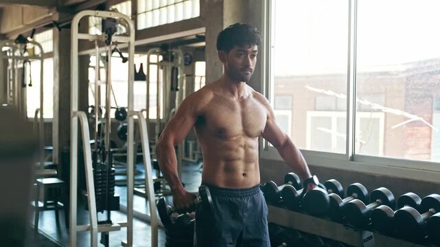 A man performs dumbbell lateral raises in a gym by a sunny window, showing defined abs and shoulders amid racks, benches, and machines.