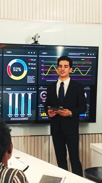 Business professional presenting data analytics on large dashboard screens during a meeting, holding a tablet while colleagues watch in a modern conference room.