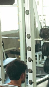 Shirtless man performing a barbell back squat in a gym rack, showcasing muscular back and shoulders among weight plates and equipment.