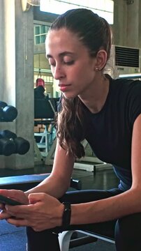 Person in gym attire resting on a bench, checking a smartphone between sets, with weights and equipment in the background.