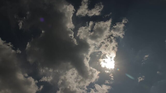 Time-lapse of bright sun shining through moving white clouds in deep blue sky with sun rays