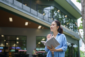 Fototapeta premium Education, university and woman in campus, Young asian woman smiles widely while tacking note. She standing in campus with her ready. Enjoying education outdoors with textbook