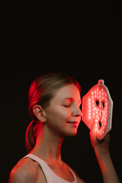 smiling woman holding infrared light therapy LED mask and looking side with eyes closed. black  background. Contrast red light. vertical composition