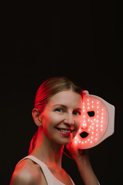 Infrared light therapy LED mask. woman holding the mask , looking to the camera and smile. black  background. Contrast red light. vertical composition