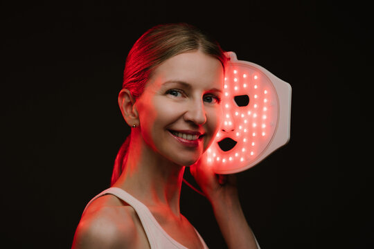 Infrared light therapy LED mask. woman holding the mask , looking to the camera and smile. black  background. Contrast red light. horizontal composition