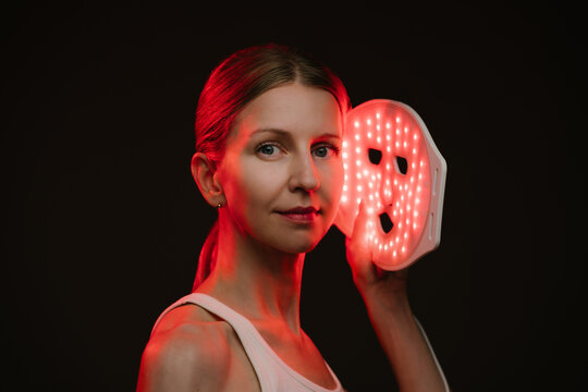 woman holding infrared light therapy mask and looking to the camera. black  background. Contrast red light