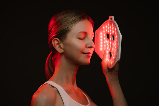 blonde woman holding infrared light therapy LED mask and looking side with eyes closed. black  background. Contrast red light. horizontal composition