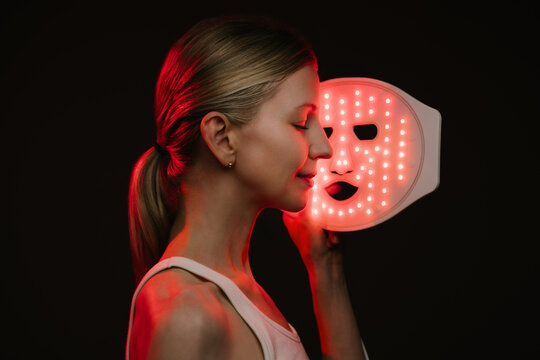 smiling woman holding infrared light therapy LED mask and looking side with eyes closed. black  background. Contrast red light. horizontal composition