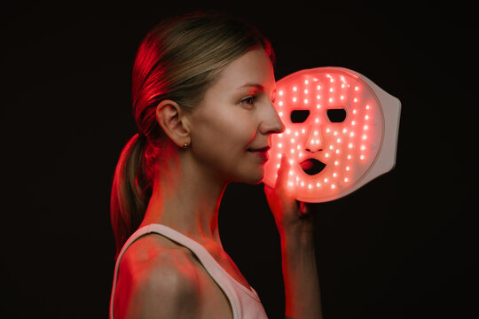 smiling woman holding infrared light therapy LED mask and looking side. black  background. Contrast red light