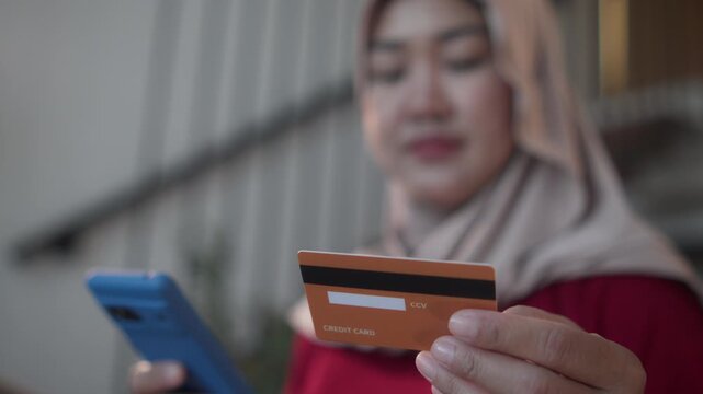 Close-up of a credit card held by a Muslim woman wearing a hijab while using a smartphone for online shopping, illustrating e-commerce, secure online payment, digital banking, and fintech.