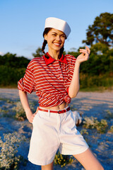 Young woman wearing sailor hat and striped shirt smiles while posing outdoors during sunset. A cheerful lifestyle lifestyle portrait capturing joy and summer fashion in a natural landscape setting.
