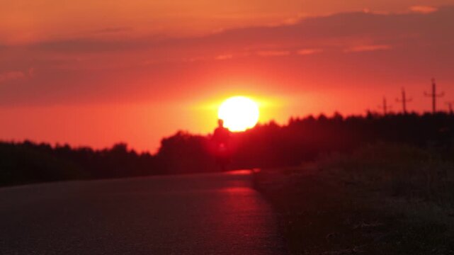 Motorcyclist riding away on a rural road toward bright orange sunset, silhouette against evening sky