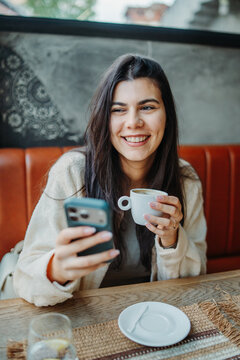 Young woman smiling while drinking coffee and using smartphone