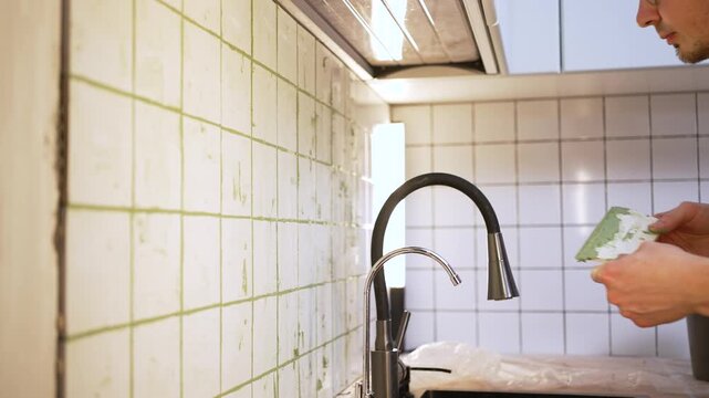 Budget-conscious young homeowner carefully applying green grout between white ceramic tiles on a kitchen wall, demonstrating diy skills during a home improvement project to save money