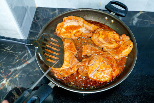 Marinated pork steaks sizzling in a frying pan with savory bubbling sauce. A black spatula flips a juicy meat cutlet on a stovetop. Home cooking, delicious dinner preparation and protein rich meal.