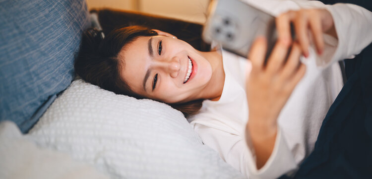 Young adult asian woman lying comfort on bed relaxing at night bedroom scrolling on phone for social media, digital dating online