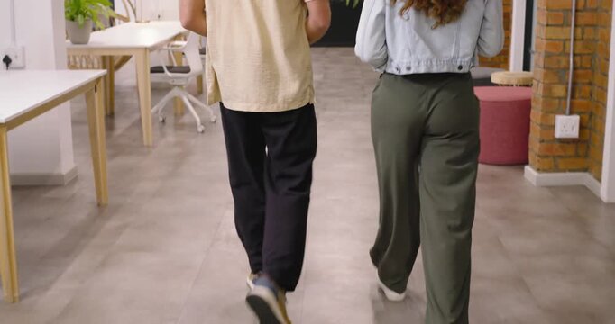 Diverse coworkers walking down office hall holding tablet, approaching planter sparking discussion