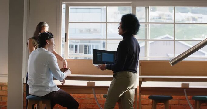 Diverse coworkers at bench by window watching man entering with laptop, placing it and demoing