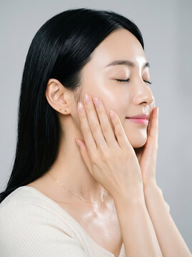 Beautiful young east asian woman with glowing skin gently touches her smooth cheeks with soft hands while posing gracefully against a clean light grey studio background